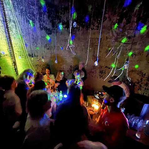 Eine Gruppe von Menschen feiert in einem stimmungsvoll beleuchteten Raum mit hängenden Glühbirnen und bunten Lichteffekten, während ein DJ Musik auflegt.