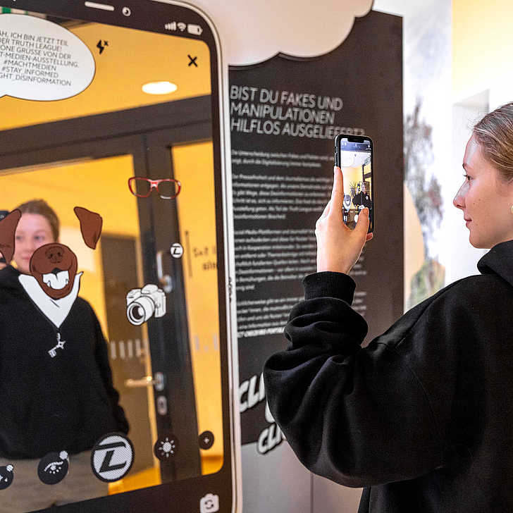Eine Besucherin der Ausstellung "Macht Medien!" zückt ihr Smartphone und richtet es auf eine Ausstellungstafel, die wie ein großes Smartphone anmutet.