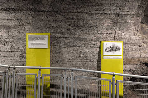 Zwei Ausstellungstafeln mit Zeitzeugenbericht und Zeichnung an einer Betonwand in der Gedenkstätte Mittelbau-Dora.