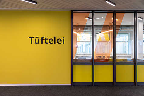 Beschreibung: Eine gelbe Wand mit der Aufschrift "Tüftelei". Große Fenster mit orangen Akzenten geben den Blick in einen dahinterliegenden Arbeitsraum frei.