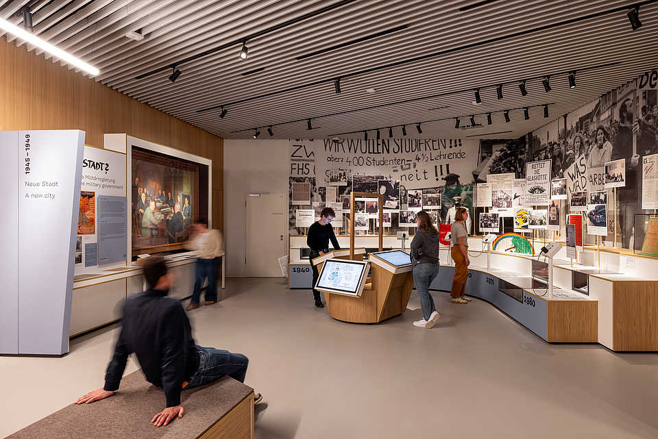 Besucher:innen in einem Ausstellungsbereich mit historischen Fotografien, Dokumenten und interaktiven Medienstationen.