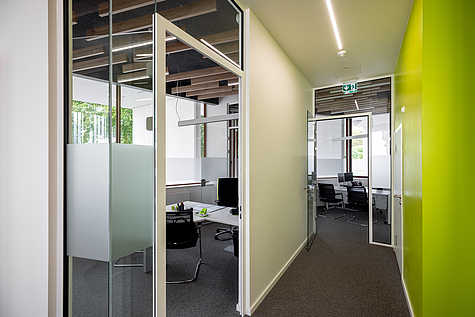 Flur mit Glastüren zu modernen Büroräumen, ausgestattet mit Bürostühlen und Monitoren. Die Decke zeigt sichtbare Holzlamellen, eine grüne Wand setzt einen farblichen Akzent.