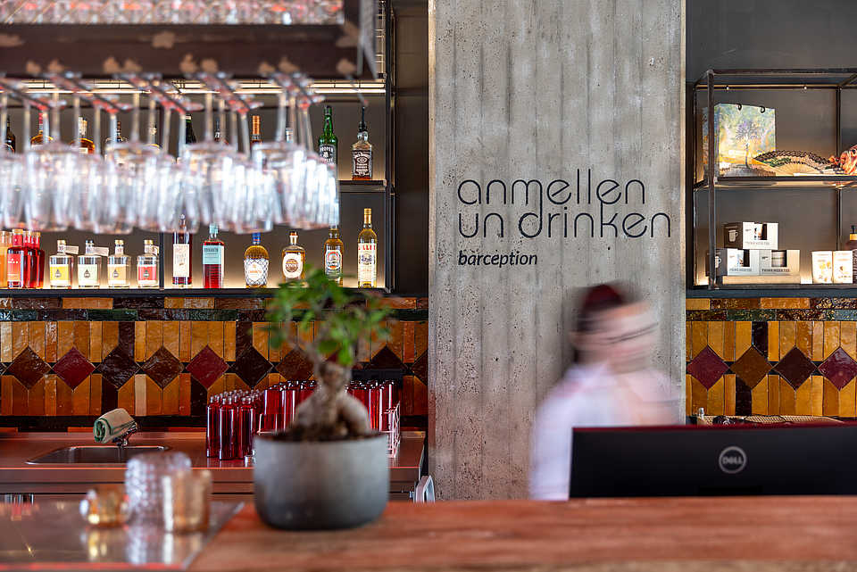 Eine Bar im industriellen Stil mit einer Wandbeschriftung „anmellen un drinken – barception“. Im Vordergrund stehen Flaschen und Gläser, während im Hintergrund eine Regalwand mit weiteren Spirituosen zu sehen ist.