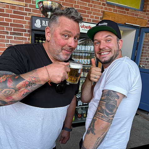 Zwei Männer posieren fröhlich für die Kamera, einer hält ein Bierglas, während der andere mit einem breiten Lächeln den Daumen hoch zeigt. Im Hintergrund ist ein Kühlschrank mit Getränken vor einer Ziegelfassade sichtbar.