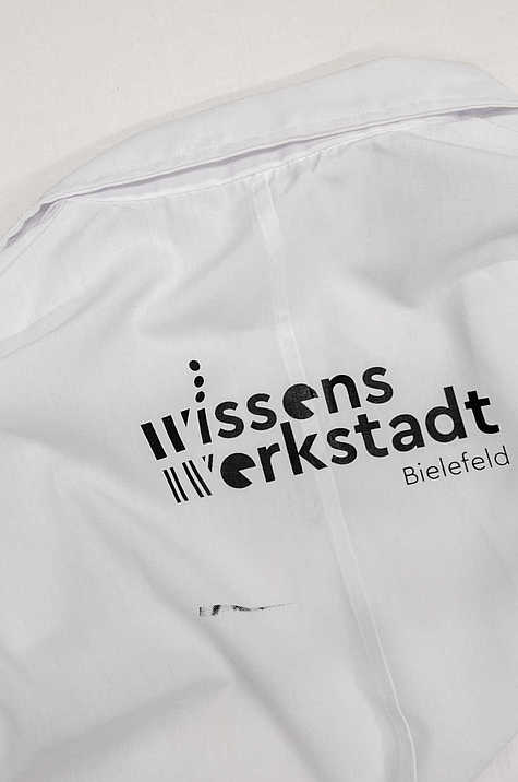 Nahaufnahme eines weißen Kittels mit dem aufgedruckten Logo der WissensWerkStadt Bielefeld.