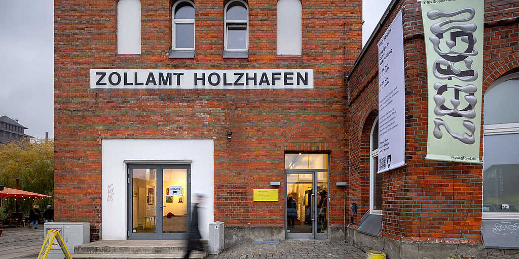 Fassade des historischen Zollamts Holzhafen mit Bannern, die die Ausstellung 'GfG Zeigen' ankündigen. Menschen gehen vorbei, während im Inneren Kunstwerke durch beleuchtete Fenster sichtbar sind.
