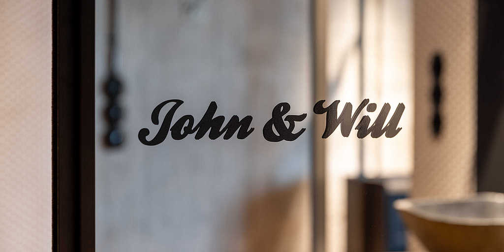 Nahaufnahme einer Glastür mit der Beschriftung „John & Will“ in stilvoller, geschwungener Schrift, die das Licht reflektiert.